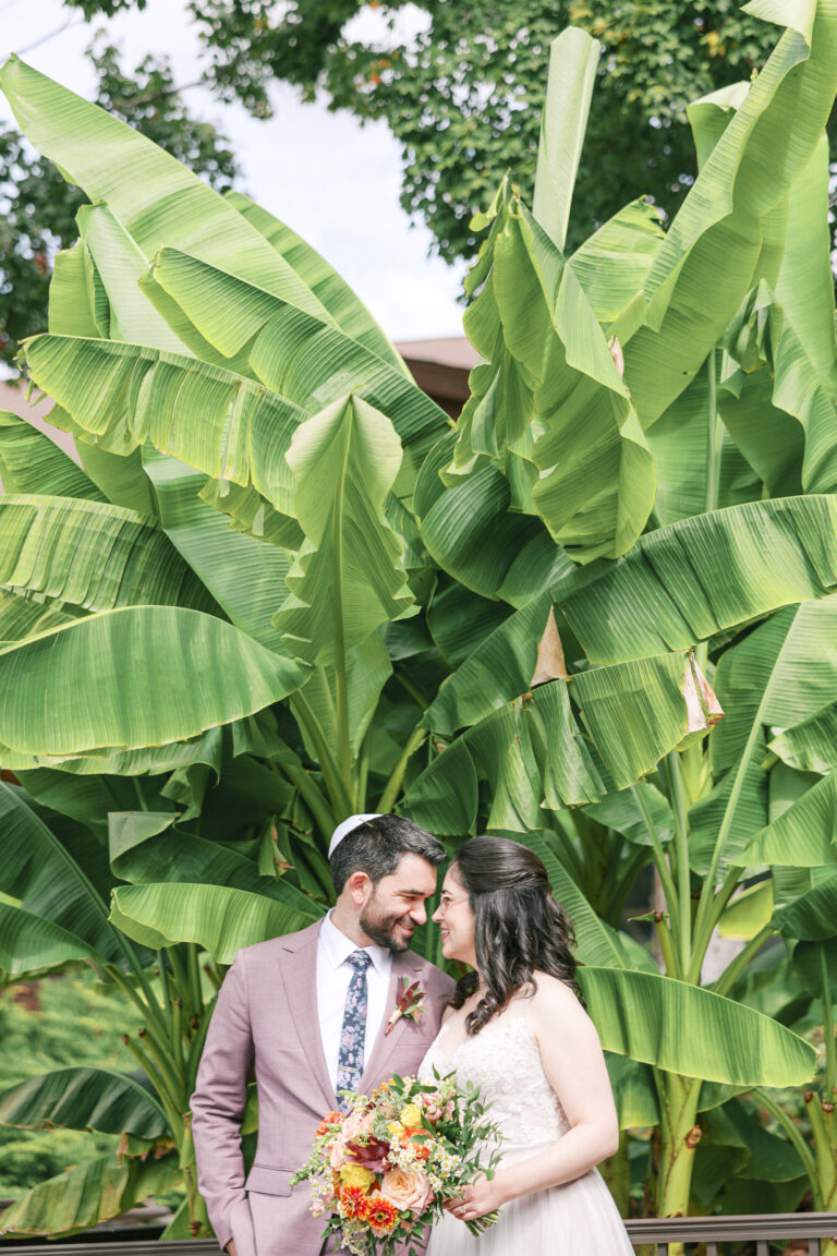 Jewish couple posing in front of a banana tree, with the groom in a light purple suit that complements the soft colors of the backdrop.