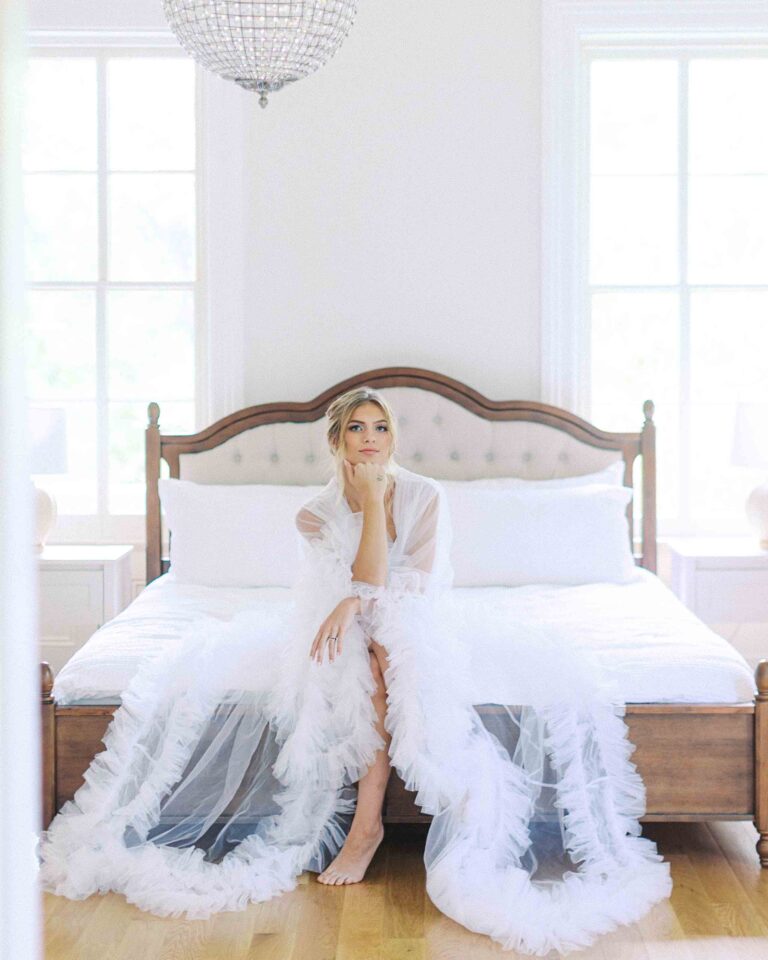 Bride seated on a white bed in a bright, elegant bridal suite, wearing a feathered robe and preparing for her wedding day.