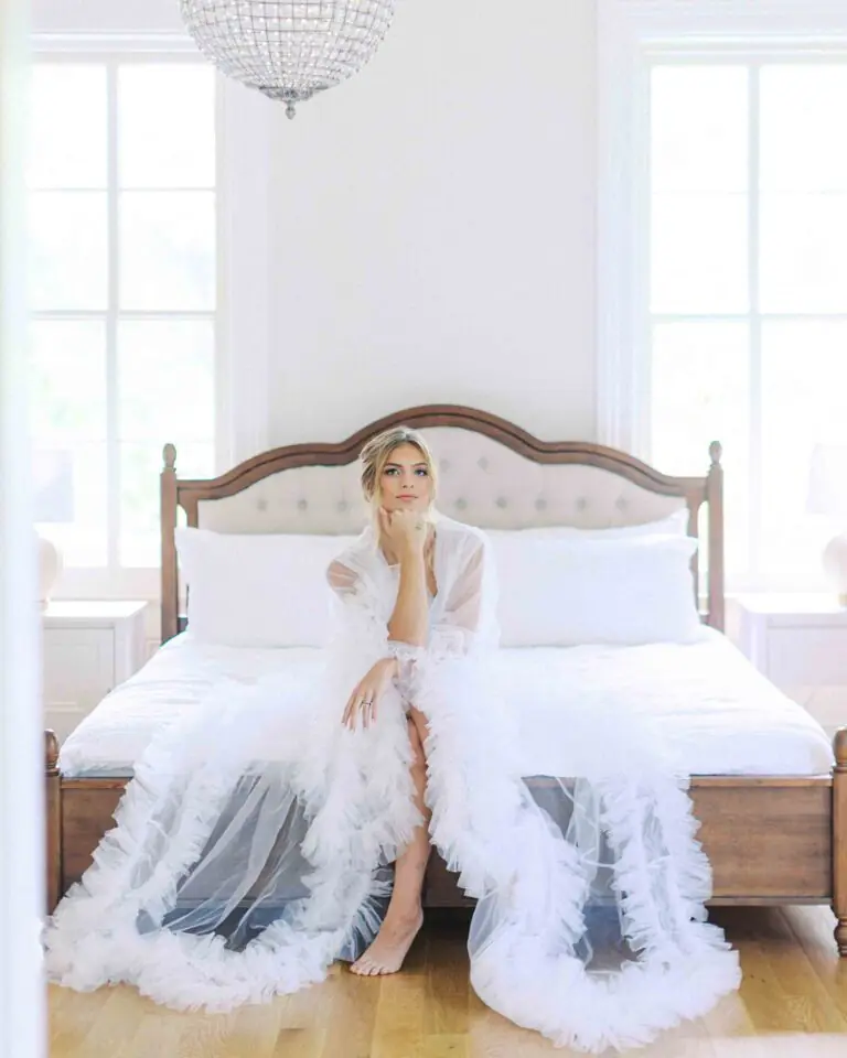 Bride seated on a white bed in a bright, elegant bridal suite, wearing a feathered robe and preparing for her wedding day.